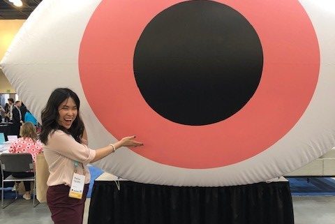 Bianca is wearing a light pink blouse and maroon-ish dress pants with black flats. She's smiling with her mouth open and eyes somewhat closed as she gestures towards a very large eye with a red pupil in the center of the convention room.