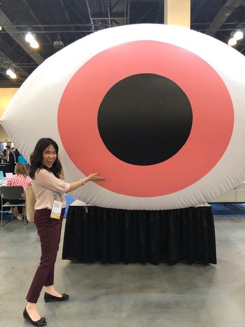 Bianca is wearing a light pink blouse and maroon-ish dress pants with black flats. She's smiling with her mouth open and eyes somewhat closed as she gestures towards a very large eye with a red pupil in the center of the convention room.