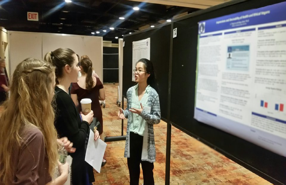 Li Ting Lin is wearing a white and gray cardigan with geometric patterns over a white dress shirt and black dress pants. The poster with her information in the background is blurred. Two students stand in front of her, one holding a coffee cup and one with a water bottle.