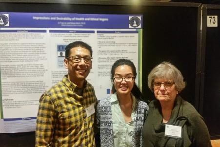 The person on Li&#039;s left is wearing a black sweater and a name tag. The person on her right is wearing a black and yellow plaid shirt. They all wear glasses. The poster behind them is, again, blurred.