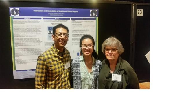 The person on Li's left is wearing a black sweater and a name tag. The person on her right is wearing a black and yellow plaid shirt. They all wear glasses. The poster behind them is, again, blurred.