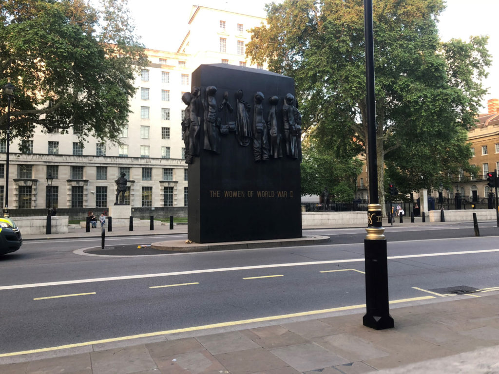 The Women of World War II monument standing in the middle of a street. The words, "The Women of World War II," are written in gold towards the bottom of the tall, rectangular block. Women's war uniforms are carved out of the stone and appear to hang off of the rectangle.
