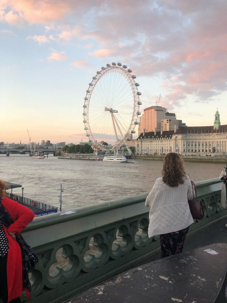 The London Eye on the River Thames. A few large buildings are behind the Ferris wheel. The photographer and a few other people stand on a bridge diagonal from the London Eye.