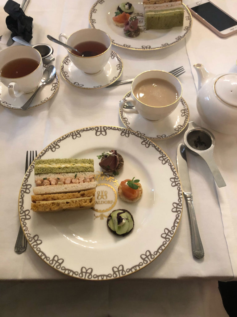 A table setting with a fork and knife on either side of the plate. There are three small sandwiches on the plate: one with presumably spinach bread, one with white bread, and one with a tan, grainy bread. There are three crackers to their right: two darker ones with meat and cucumbers respectively and a lighter cracker that appears to have salmon on top.