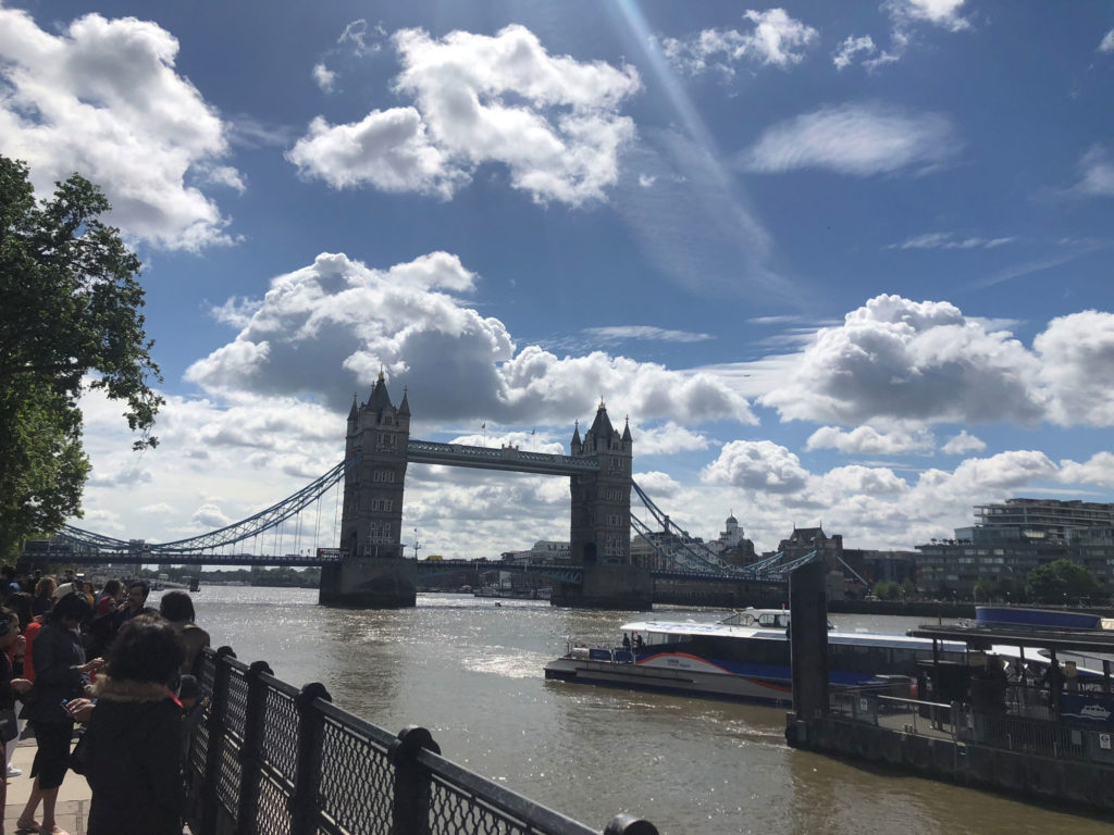 A picture from the gated walkway near the Tower Bridge in London. A ferry departs from the station in the lower right corner, and many other people stand on the sidewalk in the lower left corner. The sky is very blue and has many fluffy white clouds.