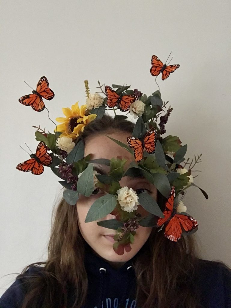 A student wears a mask made of plastic butterflies flying around leaves and flowers. You can still see the student's eyes and part of their mouth through the leaves.