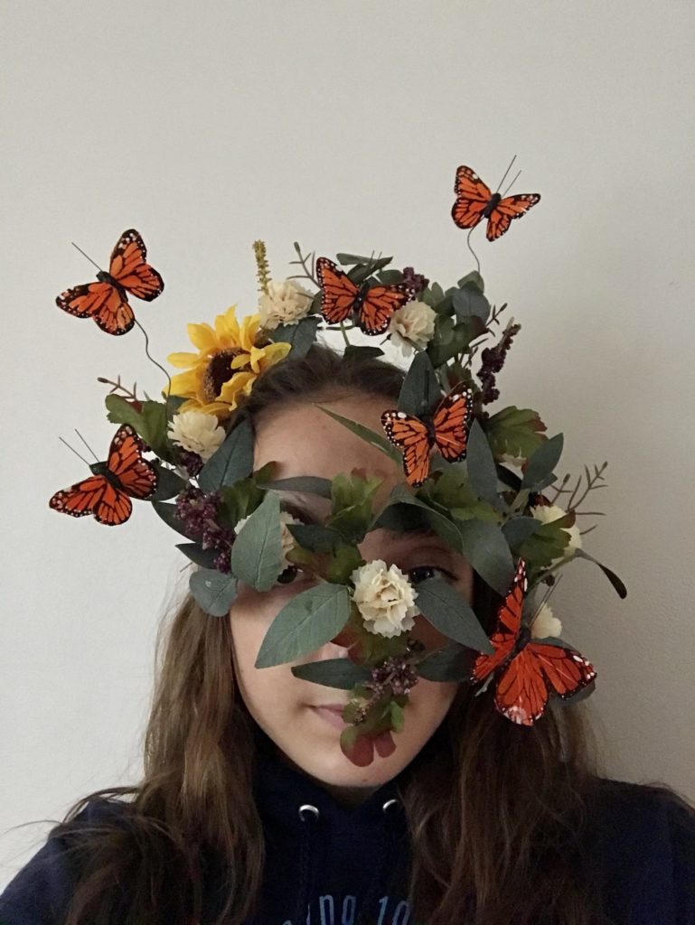 A student wears a mask made of plastic butterflies flying around leaves and flowers. You can still see the student&#039;s eyes and part of their mouth through the leaves.