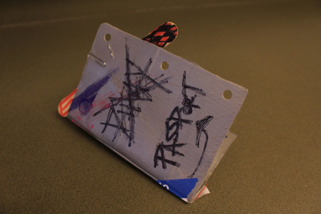 A slightly folded passport. The edge had three holes punched into it, and something is paper-clipped and stapled inside at the top right corner. A word written in marker is scribbled and crossed out. The word "passport" with an arrow underneath it is written at the bottom. A popsicle stick colored orange and black sticks out from the top.
