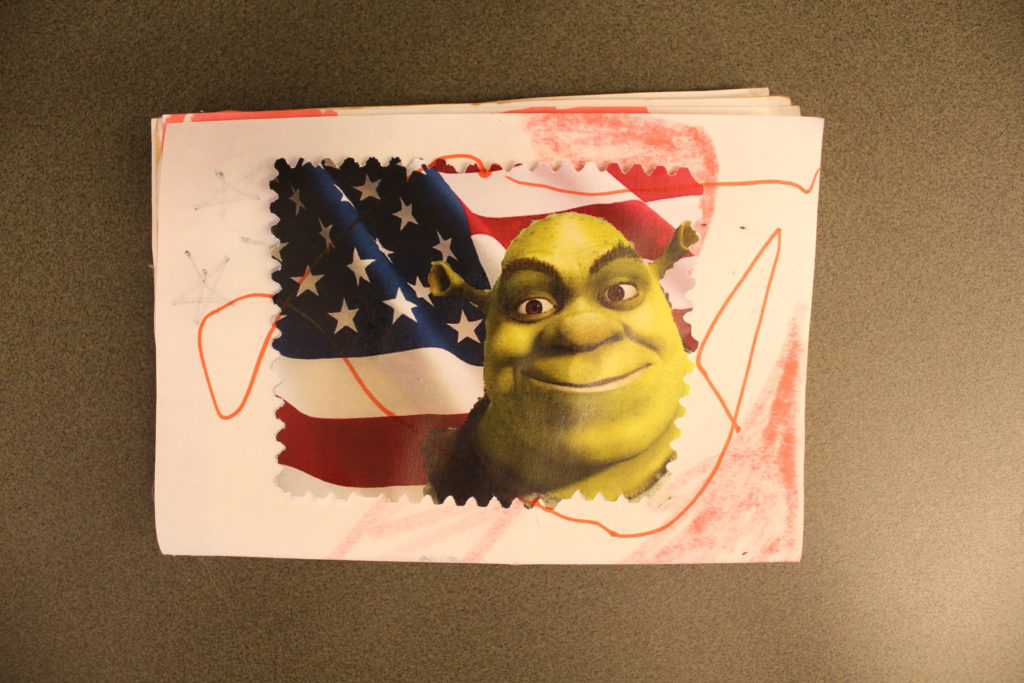 A passport made of white rectangular sheets of paper. Shrek's face is on top of a U.S. flag on a stamp. There are red scribbles on the paper.