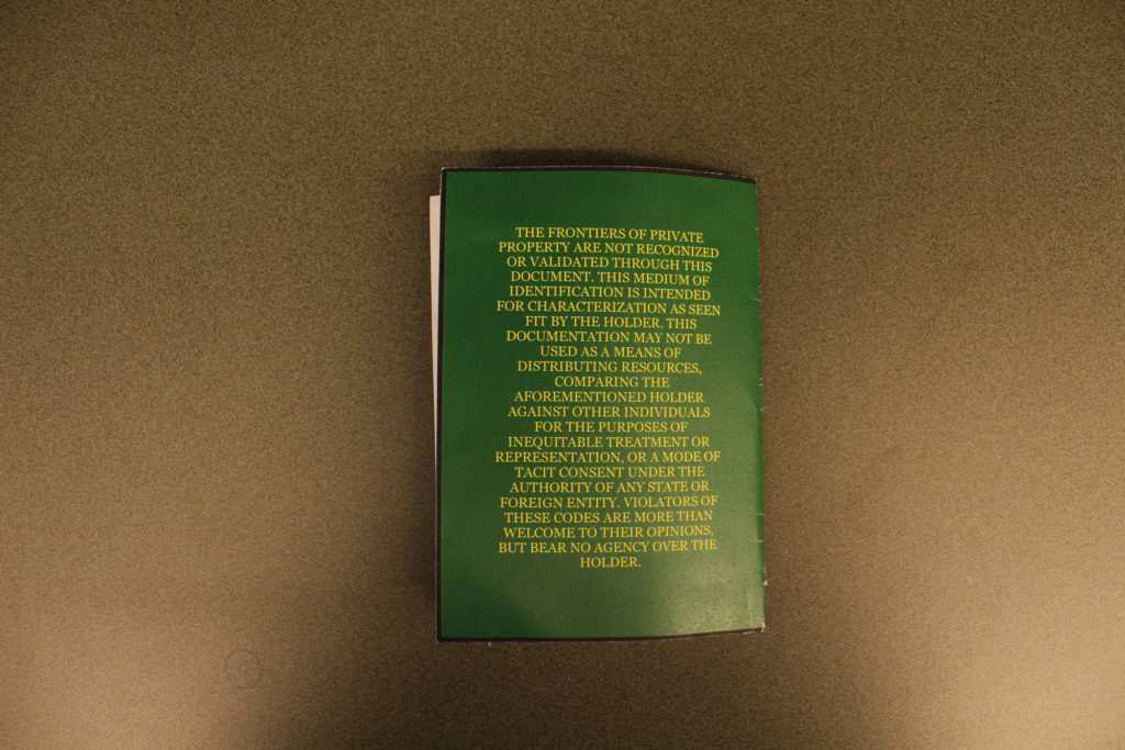The back of a green passport. The yellow centered text reads as follows: "The frontiers of private property are not recognized or validated through this document. This medium of identification is intended for characterization as seen fit by the holder. This documentation may not be used as a means of distributing resources, comparing the aforementioned holder against other individuals for the purpose of inequitable treatment or representation, or a mode of tacit consent under the authority of any state or foreign entity. Violators of these codes are more than welcome to their opinions, but bear no agency over the holder."