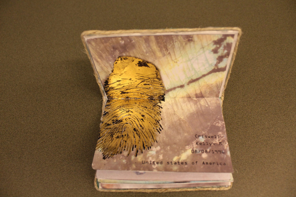 An open passport colored shades of pink and yellow pastels. A large golden fingerprint covers both pages, and the words, &quot;Criswell Kelly, M 08/08/1996 United States of America&quot; are printed on the bottom right corner.