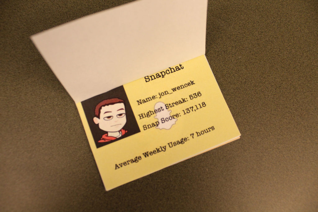 A Bitmoji, a cartoon version of a real person, is in the top left corner of the unfolded passport. They have short brown hair, tired eyes, and are wearing an orange sweatshirt. The background of the passport is yellow with the Snapchat logo. The text on the passport reads as follows: &quot;Snapchat. Name: jon_wencek. Highest Streak: 536. Snap Score: 137,118. Average Weekly Usage: 7 hours.&quot;