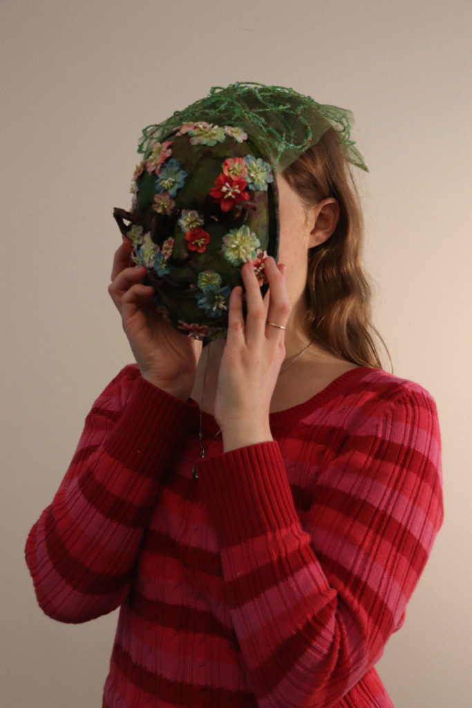 A student holds a mossy green mask over their face. It is covered with small red, blue, and pink flowers. The eye and mouth holes are very small. There is also a small piece of green see-through fabric draped over the top of the student's head like a veil.
