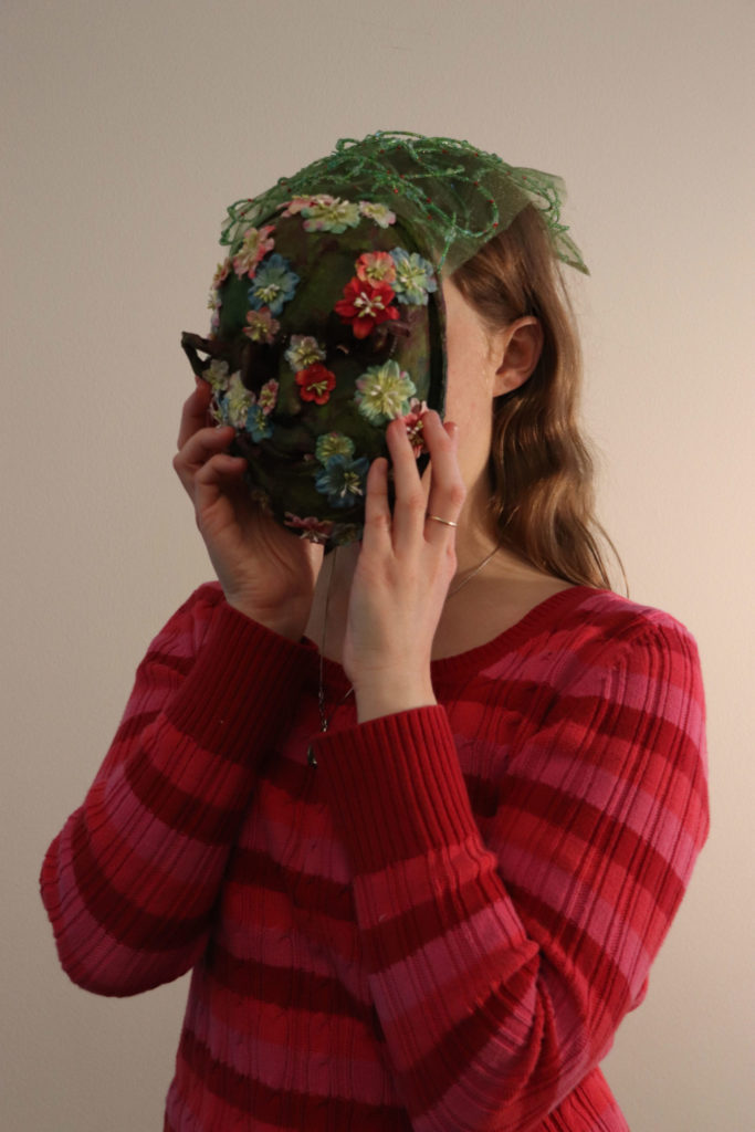 A student holds a mossy green mask over their face. It is covered with small red, blue, and pink flowers. The eye and mouth holes are very small. There is also a small piece of green see-through fabric draped over the top of the student&#039;s head like a veil.