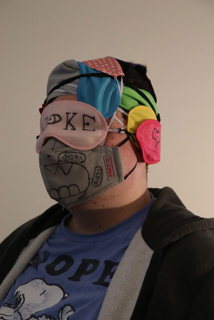 A student wearing many sleeping masks and a face mask to create one giant mask. The face mask has a large mouth with big teeth drawn on it and the word "Words" written on it in Sharpie. The mask over the student's eyes is pink and has the word "Woke" on it (the O in "woke" is an eye). The other sleeping masks on top of and around the student's head are each a different color (red, yellow, green, pink with polka dots, black, and grey).