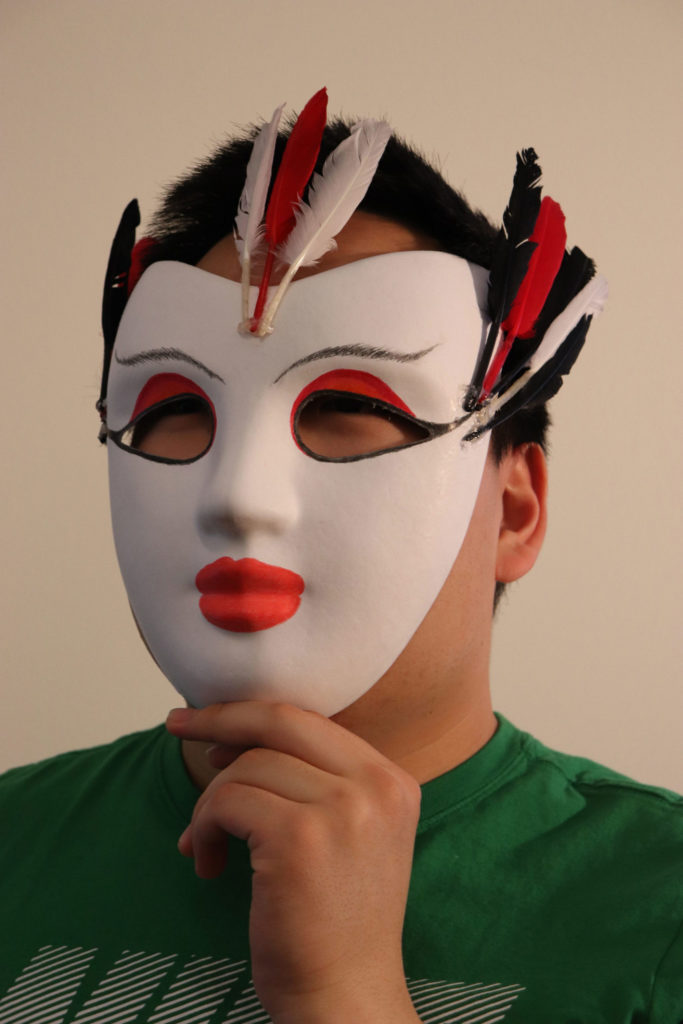 A person holding up a white mask to their face. There's red on the eyelids to mimic eyeshadow and thin, feathery brows. The lips are a bold red, and there are a few feathers each on both outer corners of the eyes and in the middle of the forehead.