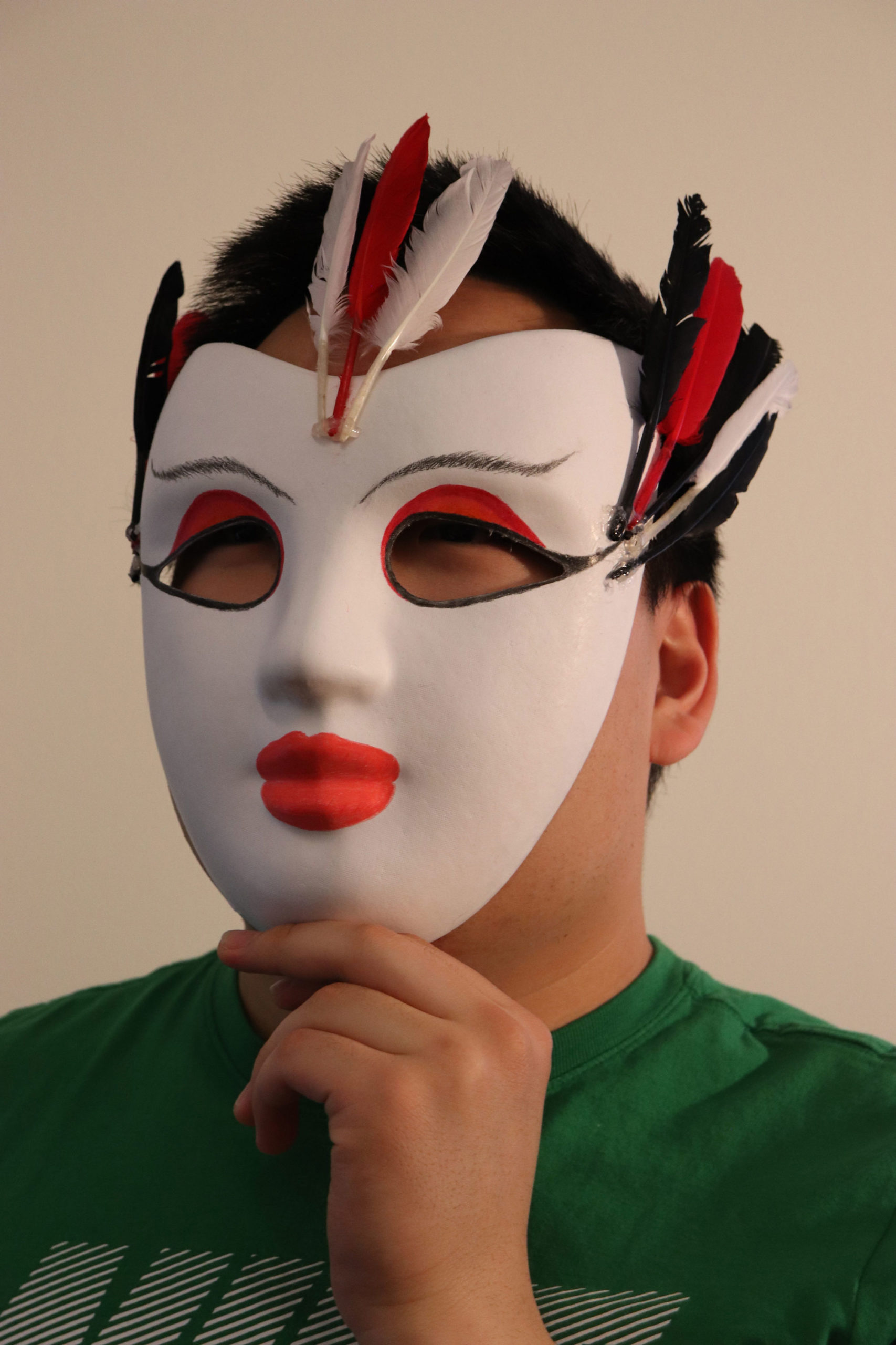 A person holding up a white mask to their face. There's red on the eyelids to mimic eyeshadow and thin, feathery brows. The lips are a bold red, and there are a few feathers each on both outer corners of the eyes and in the middle of the forehead.