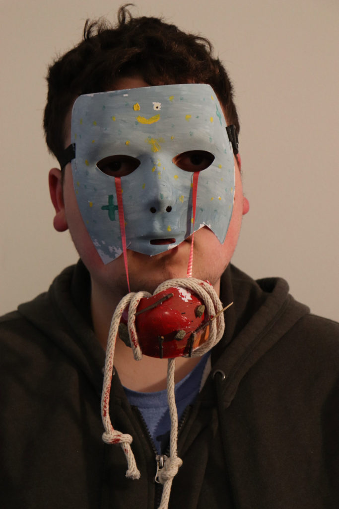 A person wears a broken blue mask on their face (secured by an elastic strap). The bottom of the mask barely covers their mouth. There are little flecks of yellow and bluish-green everywhere. A red ribbon comes out from either eye, holding up a red blob in front of the person's chin. The red blob is wrapped with rope.