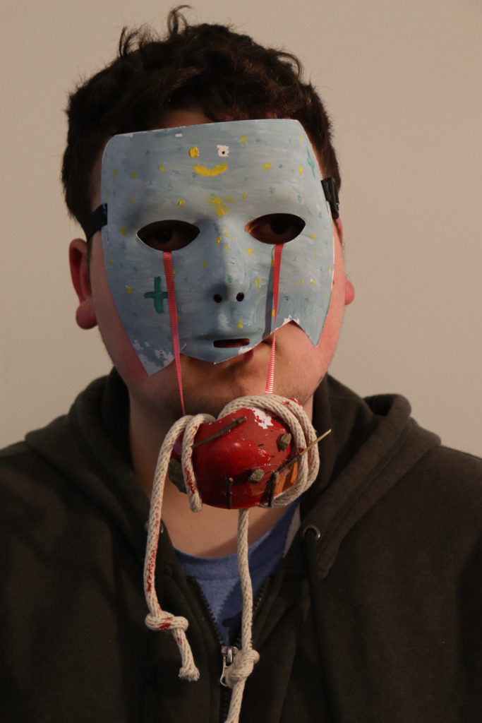 A person wears a broken blue mask on their face (secured by an elastic strap). The bottom of the mask barely covers their mouth. There are little flecks of yellow and bluish-green everywhere. A red ribbon comes out from either eye, holding up a red blob in front of the person&#039;s chin. The red blob is wrapped with rope.