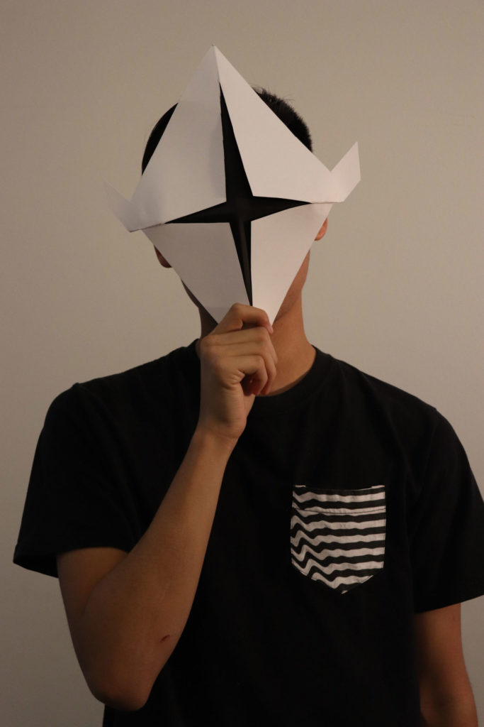 Student holding up a mask to their face. The four corners of the paper are folded in to the center, and the center of the mask is black.