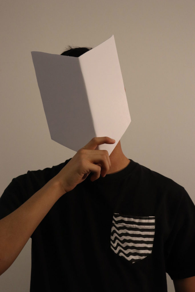 The same student from before holding up a mask that appears to be made of two parallelogram-like shapes. The left side is darker (either because it's facing away from the light or because of how it's colored), and the right side is plain white.