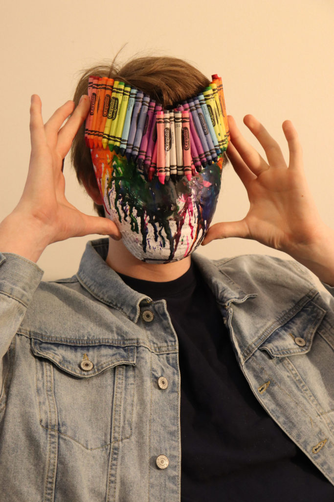 A student holds a formerly entirely white mask up to their face. A rainbow selection of Crayola crayons have been glued to the top of the mask, and their wax has dripped down to the bottom of the mask.