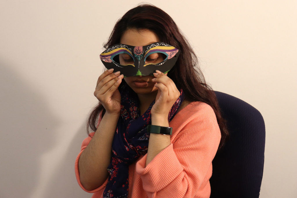 A student holds up a black Columbina-like masquerade mask. The mask covers the nose and has blue around the eyes. There's also pink, purple, and yellow lines layered on top of each other like thick eyeliner. In the center of the mask where the space between the eyebrows would be is a tiny flower drawn in a pink circle.