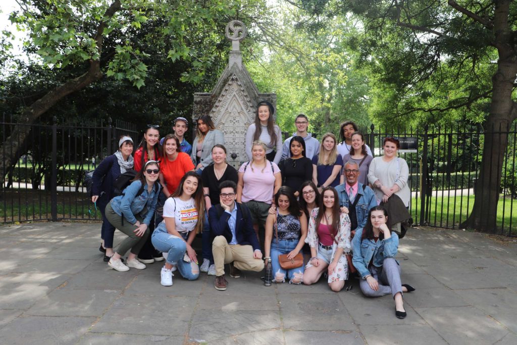 Group of students and professors standing in front of Lincoln's Inn Field. The monument behind them looks like a tiny cathedral. The peak of the building has a chimney-like stack with a circle at the top.
