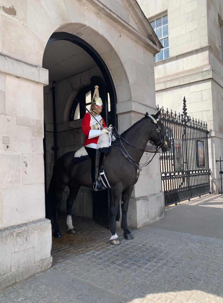 A guard wearing a red coat, long white gloves, and a gold helmet sits on a black horse. They are underneath a concrete arch, which has a black gate to its right.