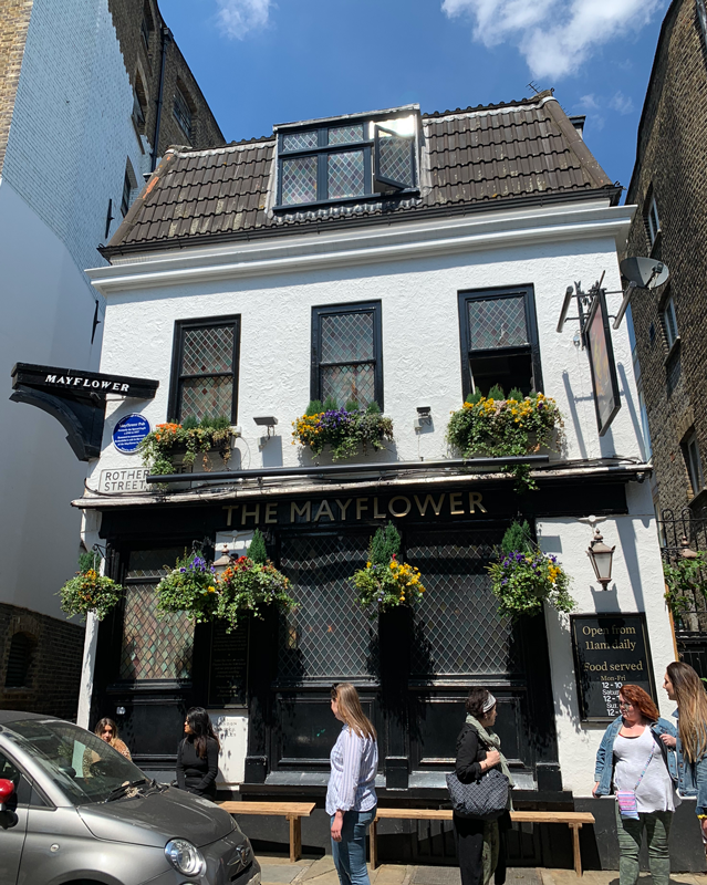 The Mayflower restaurant building. It is painted white and has three windows on the second floor, each with yellow and purple flowers under the frames. There are also flowers hanging from the sign just beneath the second floor. People stand outside the restaurant.