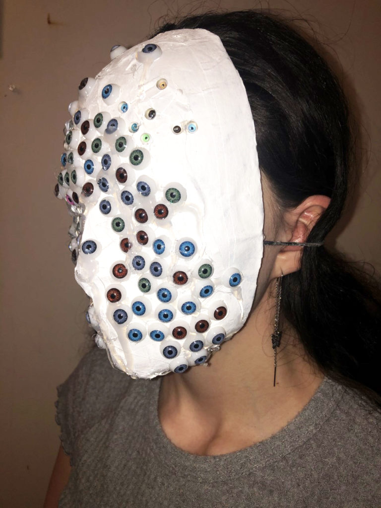 A student wearing a white mask with many fake eyes glued on it. The eyes are either blue, green, or brown.