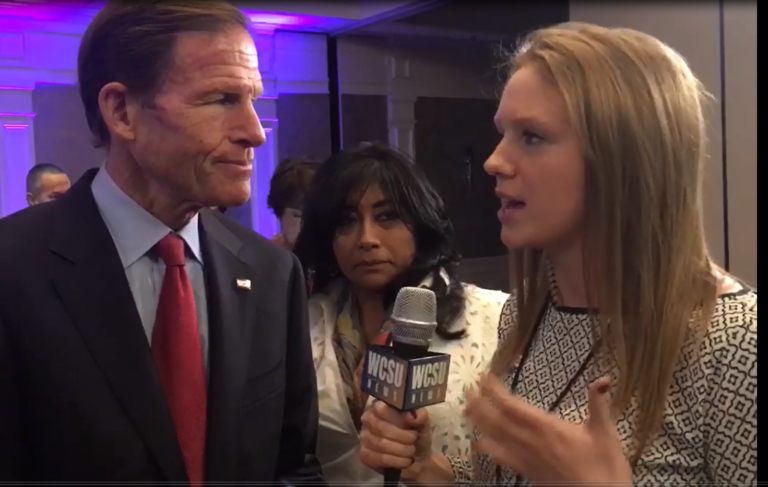 A student interviews Richard Blumenthal. Blumenthal is wearing a black jacket over a blue dress shirt. The student is wearing a white dress with a black curved diamond pattern. The student is holding a microphone.