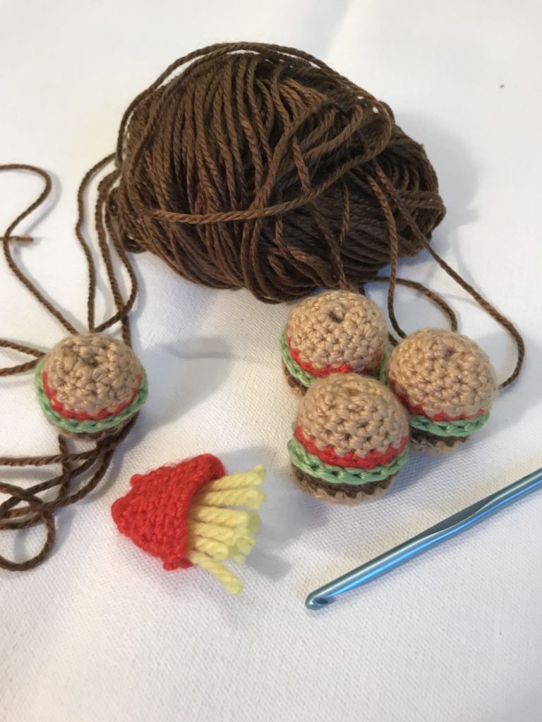 Very small crocheted burgers and french fries sit next to a small ball of brown yarn and a crochet hook.