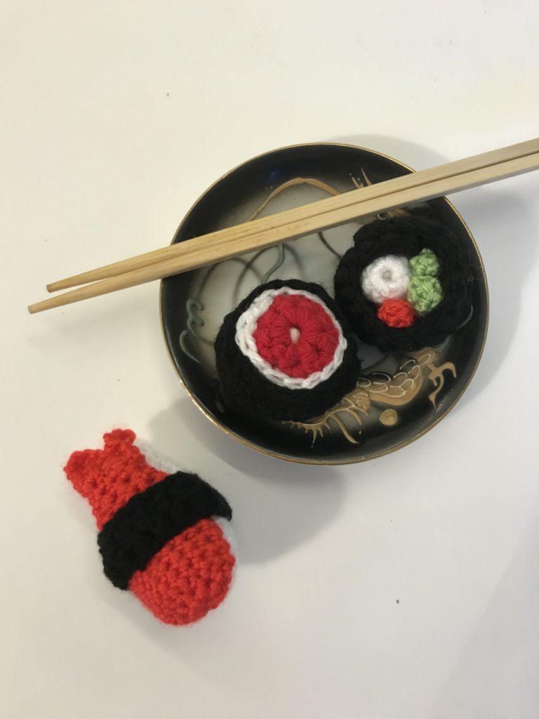 Crocheted sushi roll sits in a bowl with chopsticks resting on the rim. A piece of sushi also sits outside the bowl.