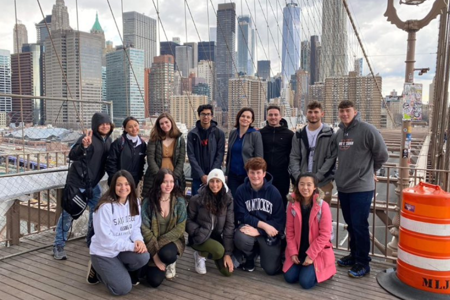 Honors students on the Brooklyn Bridge. There are eight students in the back standing and four students and the assistant director in the front kneeling. They are all wearing jackets and sweatshirts and pants.