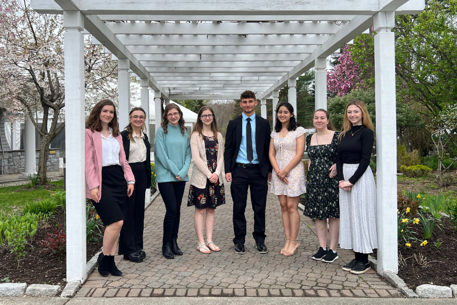 Students at Student Success Breakfast standing outside of the Amber Room in Danbury, CT. There are eight students standing in a half circle under a long trellis with flowers and trees on either side.