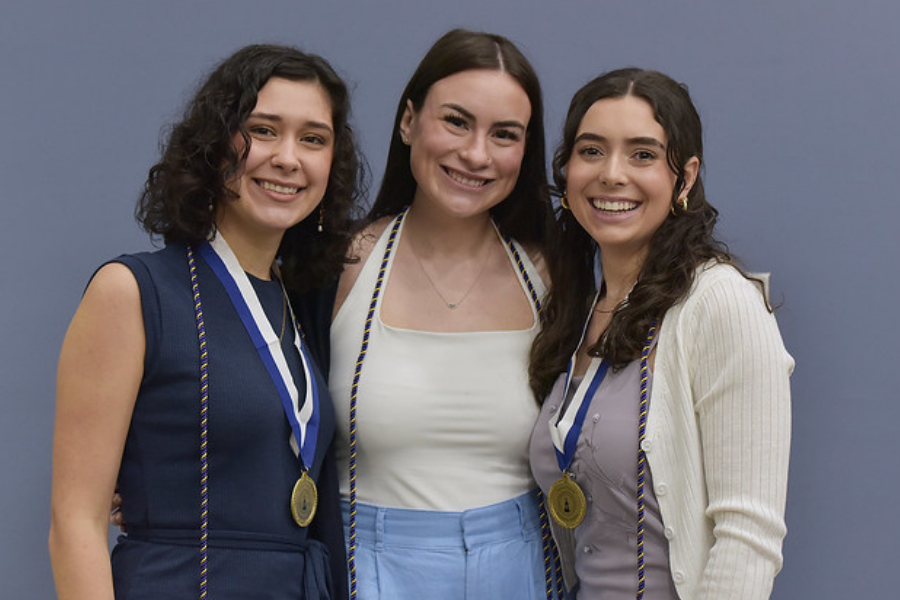 Three honors graduates standing in front of a bluish-gray background. The student in the middle has a blue and orange cord around their neck. The students on the left and right wear the cord and a gold medal with the Kathwari Honors Program logo (the tower on top of the honors building).