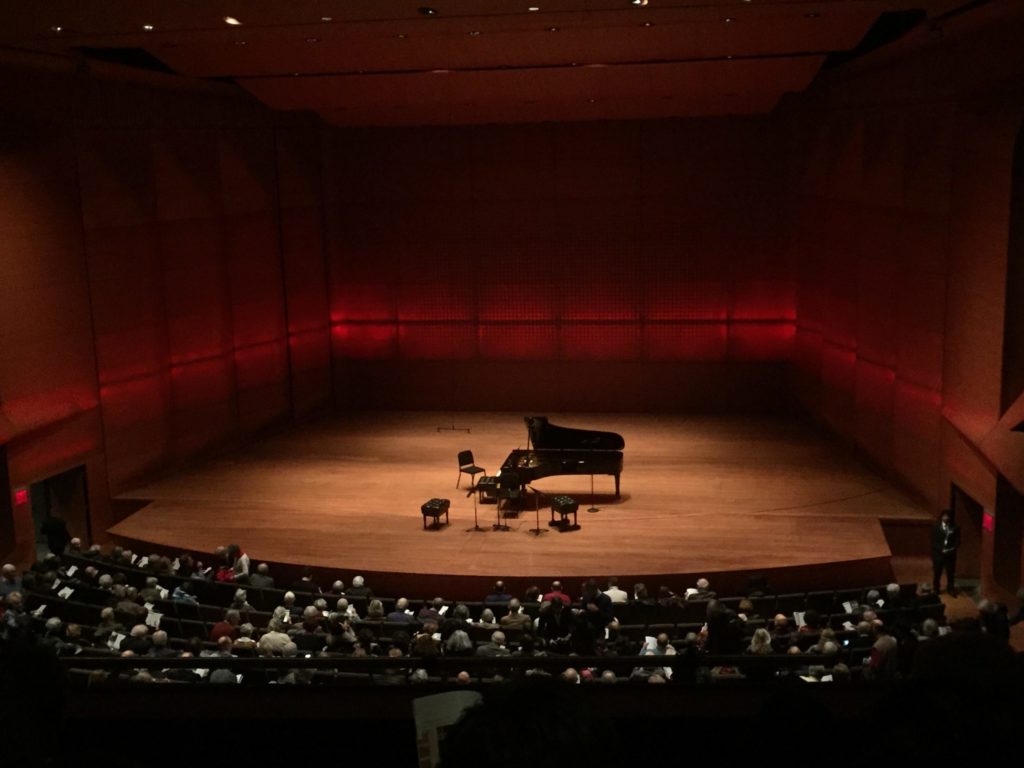 The stage at the Lincoln Center. A grand piano and empty chairs and stools are in the center.