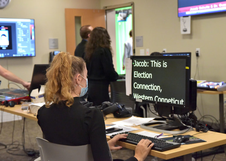 A student sits at a desktop operating the newsroom teleprompter, which says, "Jacob: This is Election Connection, Western Connecticut." The student is wearing a black long-sleeved turtleneck and silver hoop earrings.