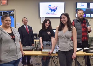 Students and faculty stand around audio equipment. The two people in front are wearing gray shirts. The person on the left in the back wears a black suit. The person in the middle wears a black T-shirt and jeans. The person on the right wears a plaid red shirt under a puffy gray vest and jeans.