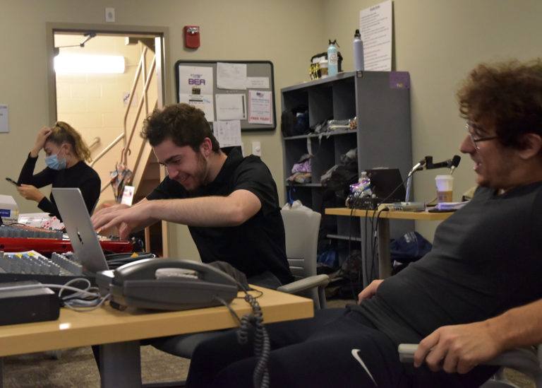 Three students sitting at a table. The student on the left wears a black long-sleeved shirt and is looking at their phone. The student in the middle is wearing a black T-shirt and gray pants and is fixing a soundboard. The student on the right is wearing a gray shirt and black pants and is leaning back in a chair.