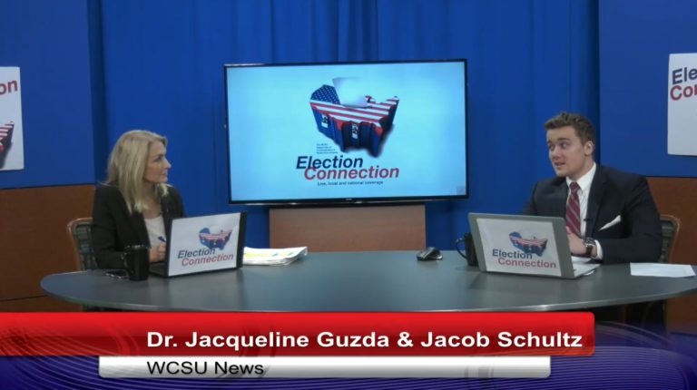 Dr. Guzda sits on the left and is wearing a black jacket over a white shirt. Jacob Schultz is on the right and is wearing a black jacket over a white dress shirt. They both have laptops in front of them which say "Election Connection" on their backs.