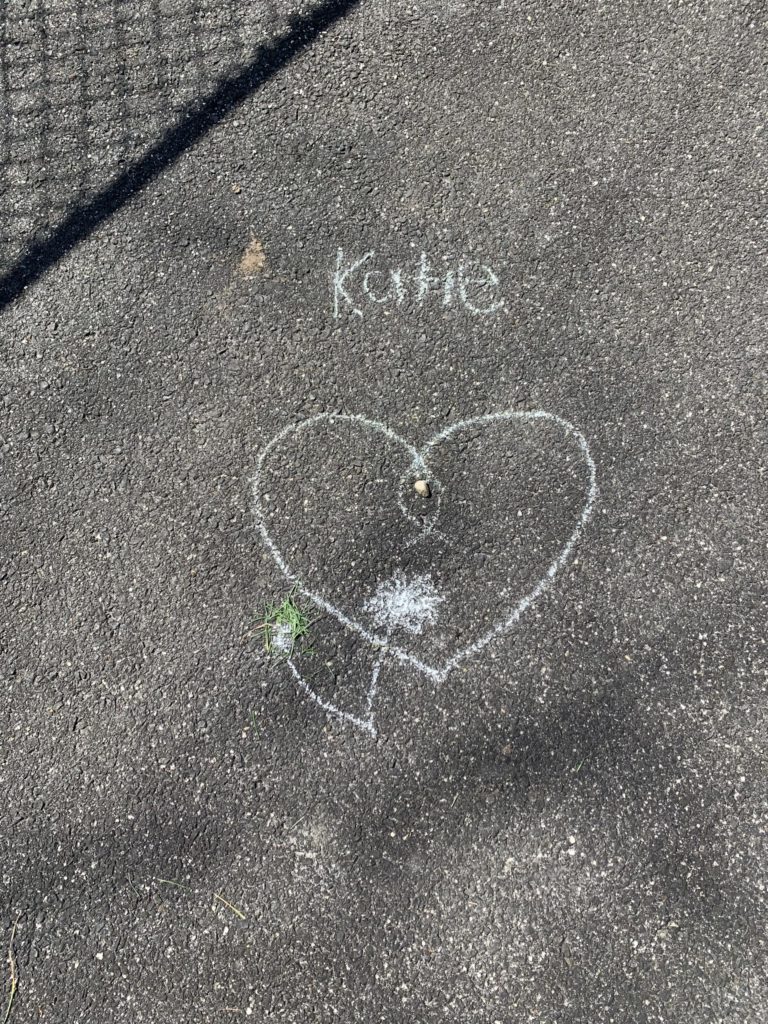A heart drawn with chalk. A tiny flower was drawn underneath it, and someone put grass clippings on the flower's stem. "Katie" is written above the heart.