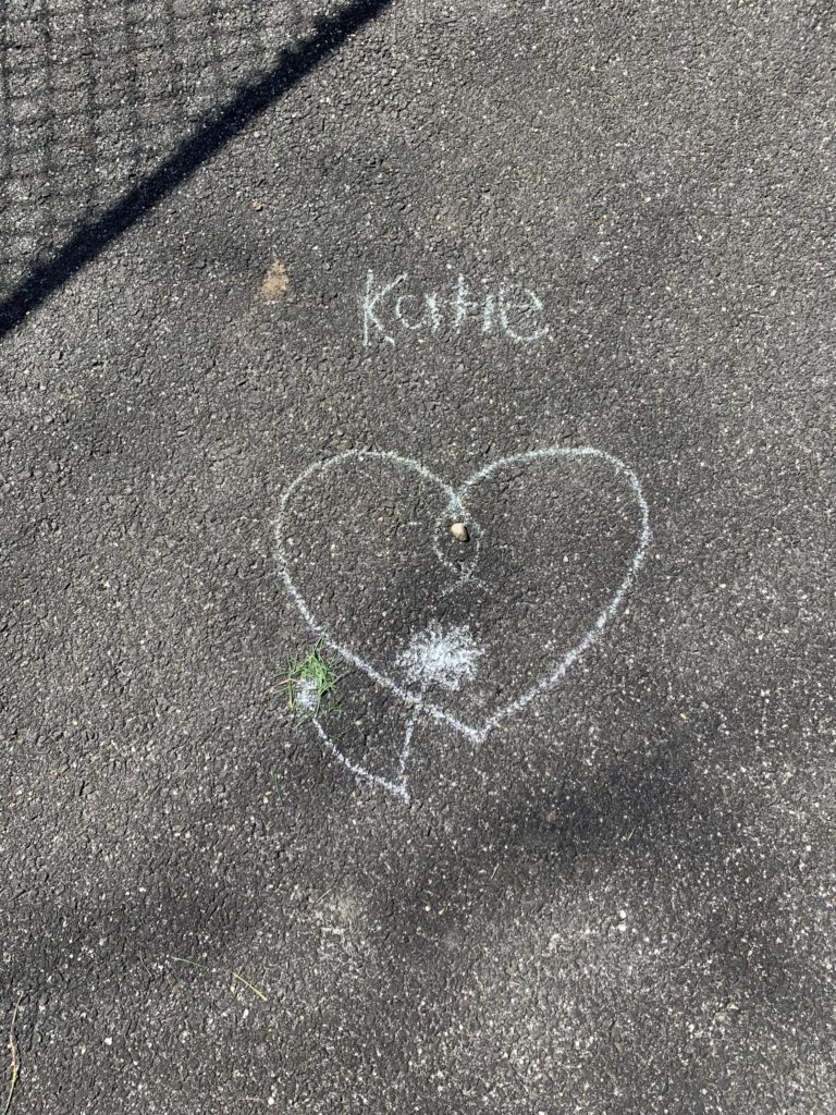 A heart drawn with chalk. A tiny flower was drawn underneath it, and someone put grass clippings on the flower&#039;s stem. &quot;Katie&quot; is written above the heart.