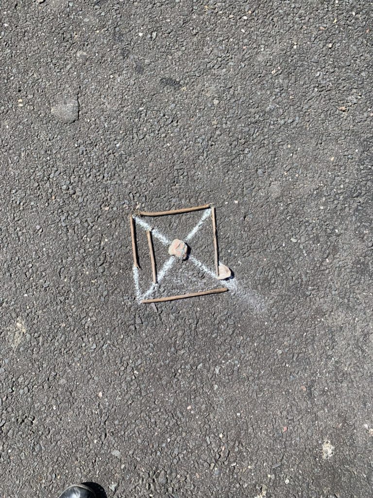 An X drawn with chalk. A rock sits in the center of the X, and a few twigs create a box around the X's edges.