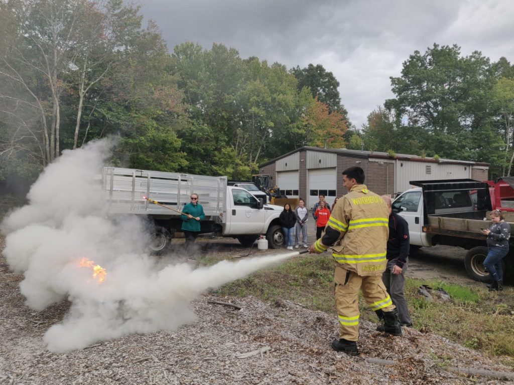 Firefighter showing students how to put out a fire with a fire extinguisher. The fire is on the left and is engulfed by the spray from the extinguisher. Students watch in the background near a couple of trucks and a garage.
