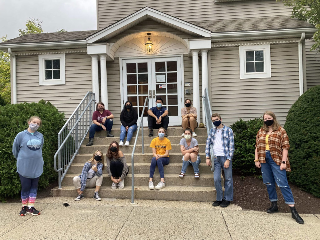 Eleven students sit on and stand near the steps of the Honors House. They are all wearing masks. There are large green shrubs on either side of the steps.