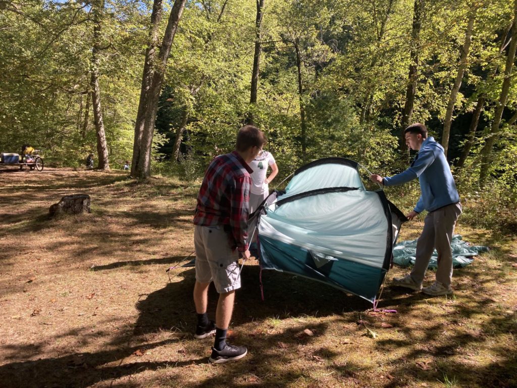 Three students setting up a blue, circular tent. They are standing on dirt covered in very little grass. There's a blanket behind tent and many trees with leaves behind them.