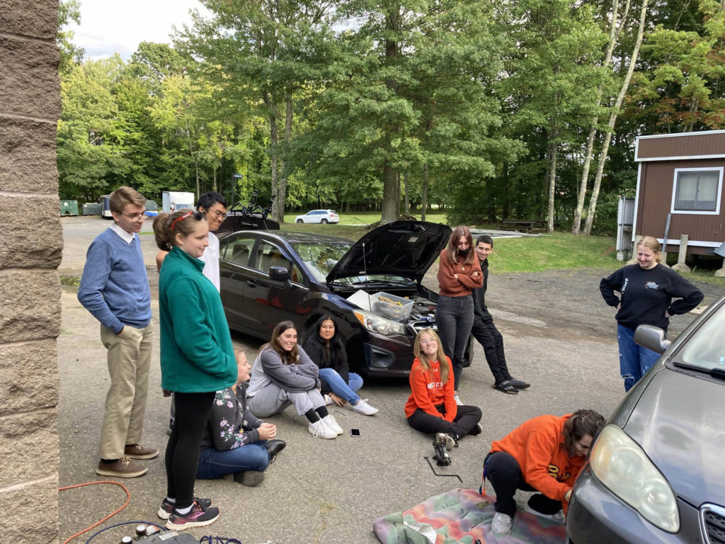 Eleven students sit and stand around two cars. The car in the background has its hood open. One student crouches near the car in the front and is measuring something on the front right tire.