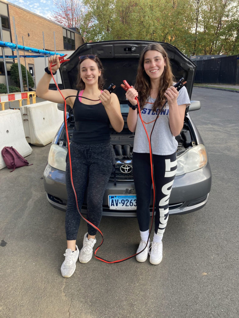 Two students standing in front of a car with an open hood. They are holding jumper cables in both hands. They have both long, brown hair and white sneakers.